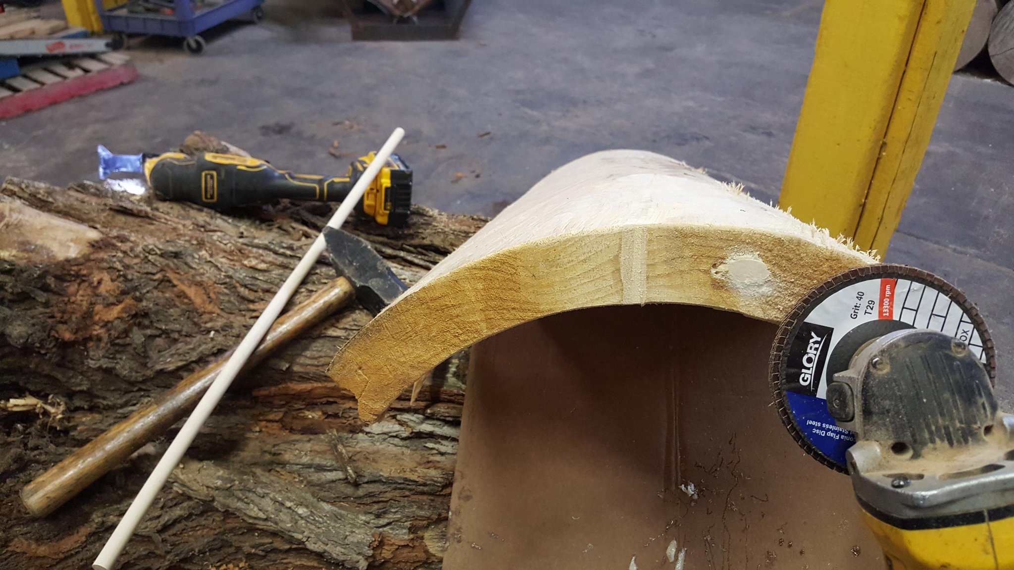 Angle grinder with Glory 40-grit flap disc smoothing the spline channel area on a bark-free IC WOOD half-log — hammer and DeWalt drill visible on the bark log behind it, IC WOOD production shop